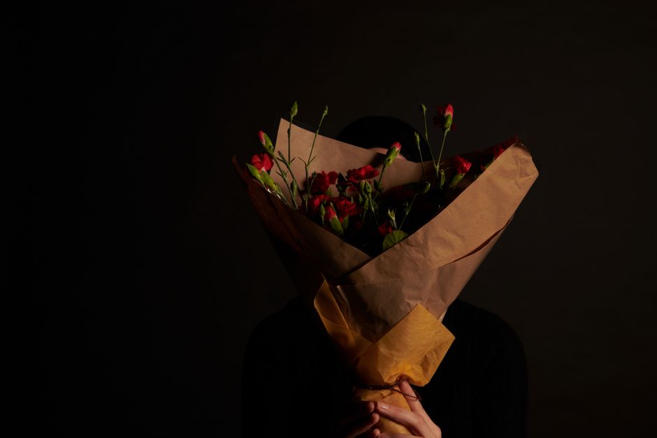 A person holding a bouquet of fresh red and pink carnations wrapped in brown craft paper with some green stems and buds visible. The bouquet is held in front of their face, obscuring their identity, w