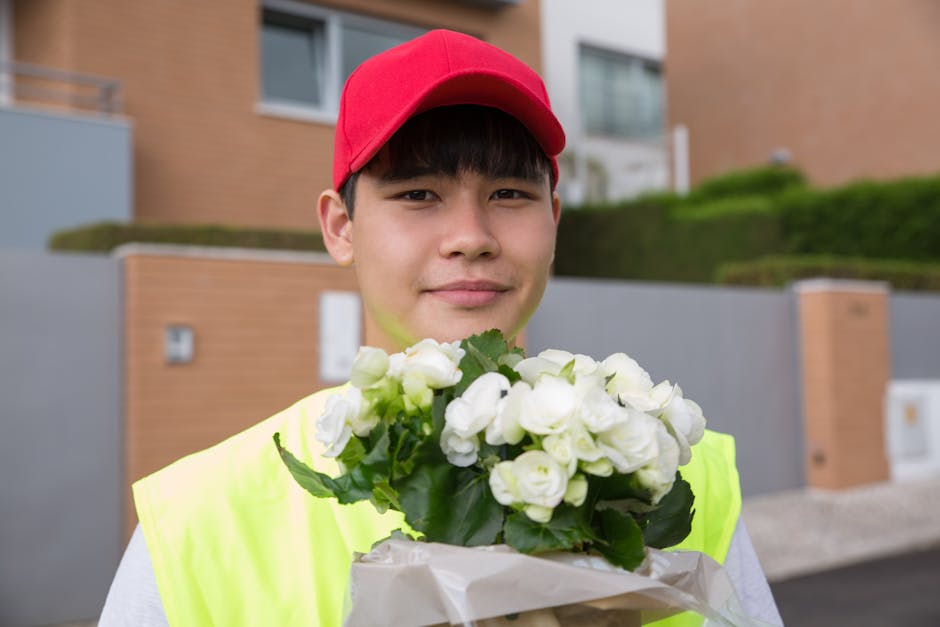 A young male delivery person with light skin, dark hair, and a friendly expression, wearing a bright yellow high-visibility vest and a red cap, holding a bouquet of fresh white flowers with green foli