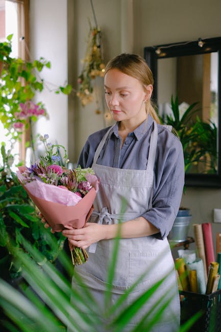 A young woman with light brown hair, dressed in a blue shirt and light gray apron, is holding a bouquet of fresh flowers wrapped in pink tissue paper. The arrangement includes vibrant purple, pink, an