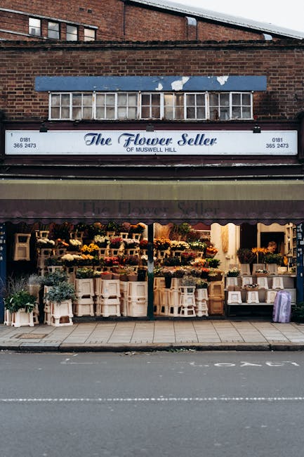 The exterior of The Flower Seller of Muswell Hill, a flower shop with a large blue and white sign above the entrance. The shop front is open, displaying a variety of fresh flowers arranged in baskets,