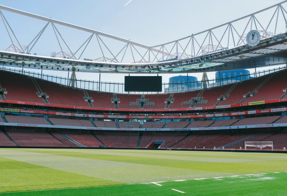 An empty football stadium with a vibrant green pitch and surrounding red seating sections. The stadium features modern architecture with a large, white, lattice-style roof structure overhead, partiall