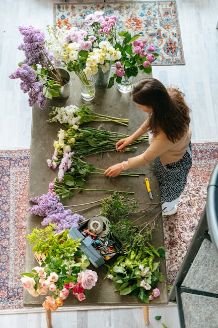 A woman arranging a variety of fresh flowers on a wooden table indoors. The arrangement includes pink and white roses, purple liatris, and other seasonal blooms, all with vibrant petals and green foli