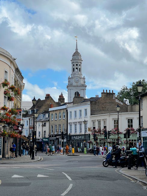 This image shows a busy street scene near Highbury Fields N5 with a prominent white clock tower featuring a golden weather vane at the top, set against a partly cloudy sky. Surrounding the tower are c