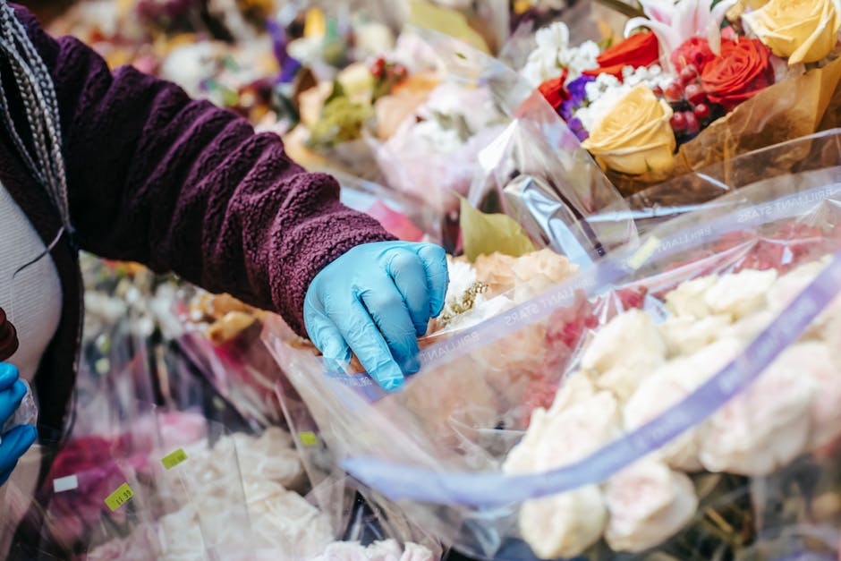 A florist wearing a burgundy textured sweater and blue gloves is selecting fresh flowers from a large display at a floral shop. The arrangement includes various roses in shades of yellow, pink, and cr