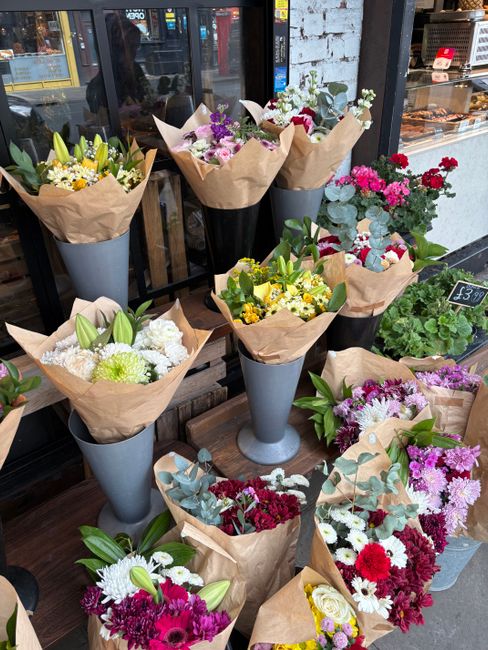 A display of various floral arrangements arranged in tall, tapered grey vases and wrapped bouquets placed on a wooden surface outside a flower shop. The flowers showcase a vibrant mix of colours, incl